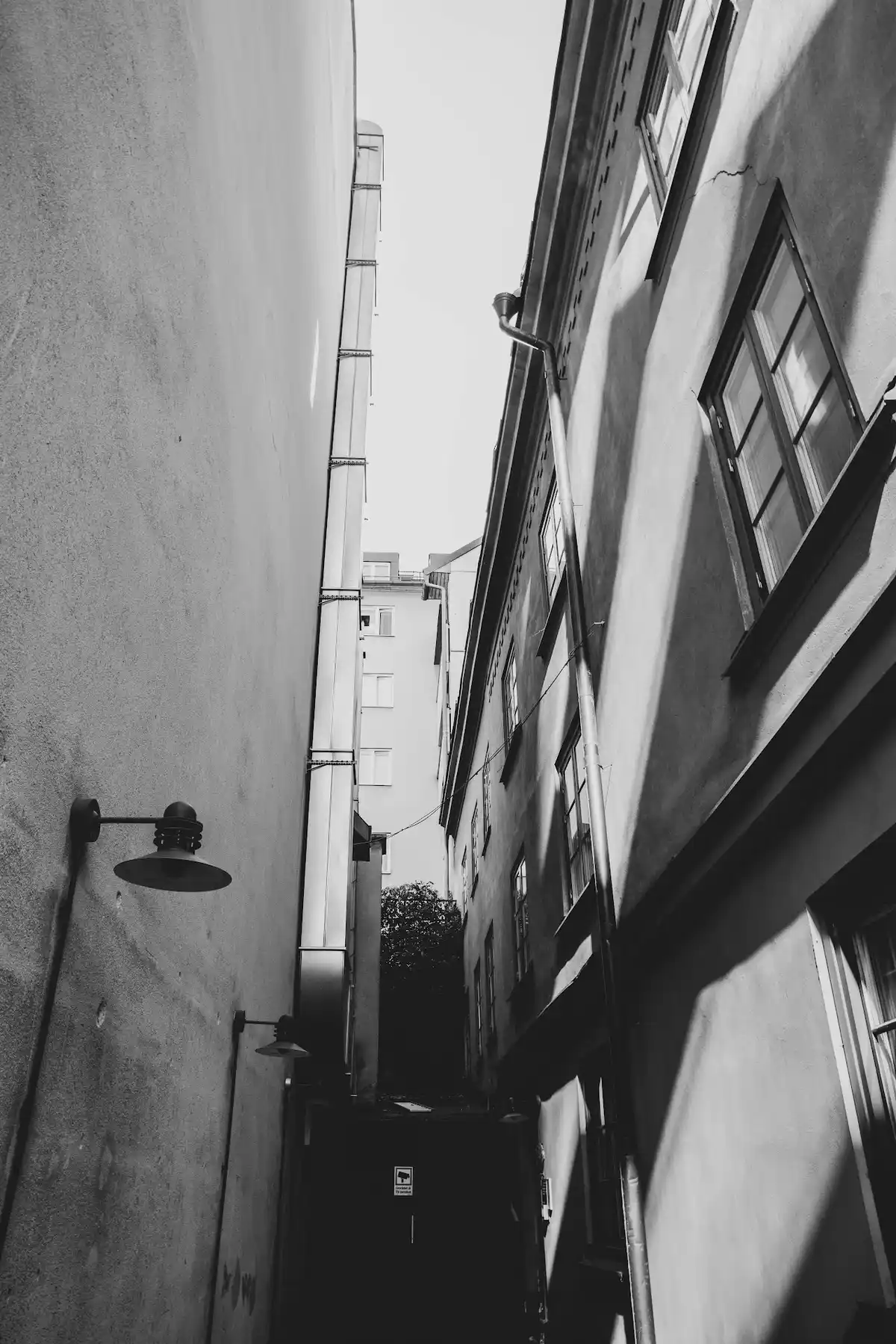 Black-and-white view through a narrow courtyard between tall walls, with lamps, pipes, windows, and daylight at the far end