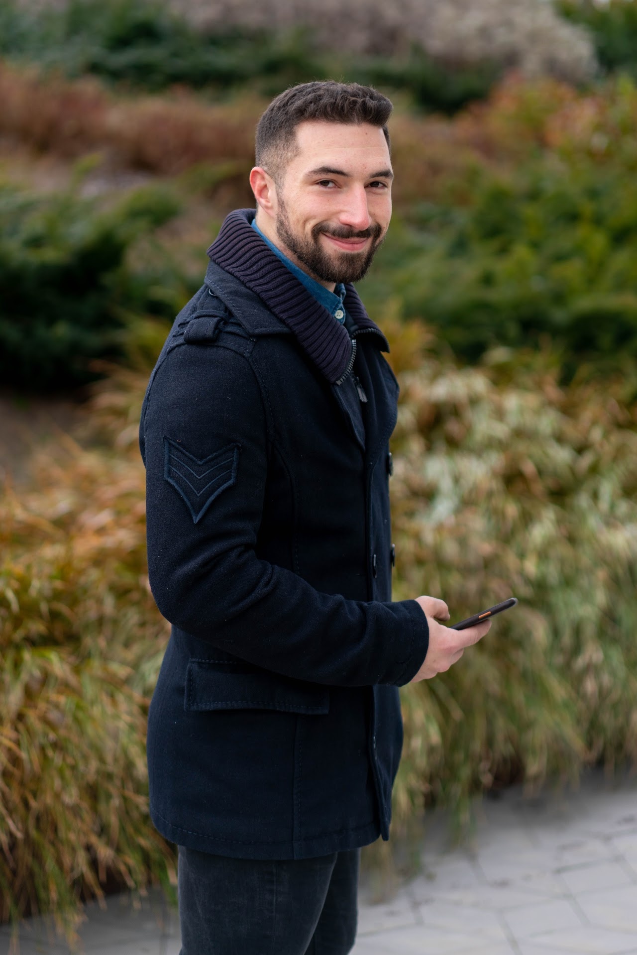 Man in a dark coat smiling outdoors by greenery